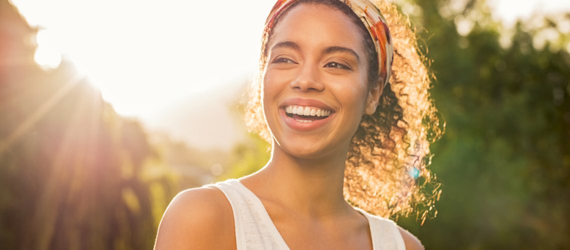 Portrait of beautiful african american woman smiling and looking away at park during sunset. Outdoor portrait of a smiling black girl. Happy cheerful girl laughing at park with colored hair band.