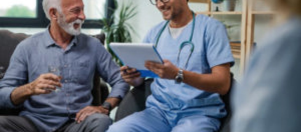 Happy senior man talking with a doctor who is reading medical reports during home visit.