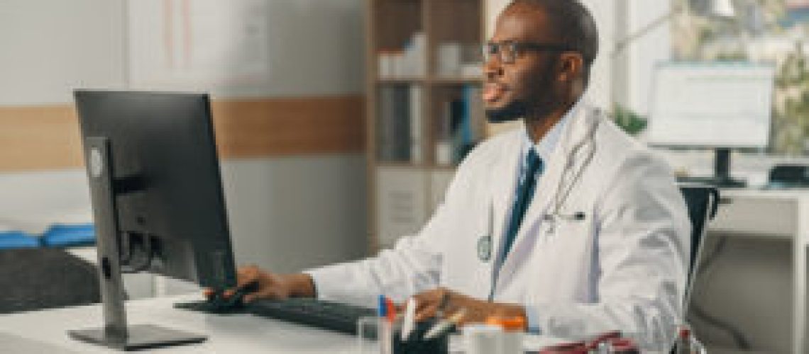 Experienced African American Male Doctor Wearing White Coat Working on Personal Computer at His Office. Medical Health Care Professional Working with Test Results, Patient Treatment Planning.