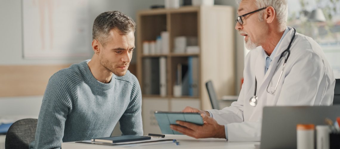 Experienced Middle Aged Family Doctor Showing Analysis Results on Tablet Computer to Male Patient During Consultation in a Health Clinic. Physician Sitting Behind a Desk in Hospital Office.